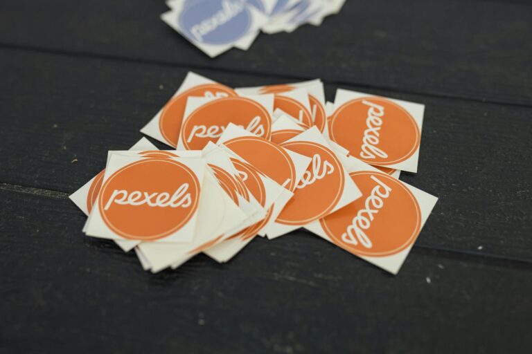 A pile of Pexels logo stickers in orange and blue on a dark wooden table.